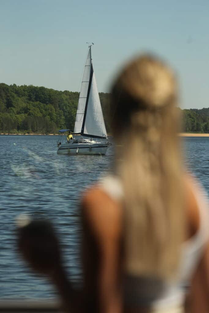 Frau blickt auf Segelboot auf ruhigem See, Natur im Hintergrund.