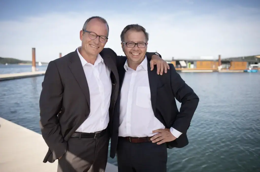 Zwei Männer in Anzügen am Wasser mit Eco Lodges im Hintergrund.