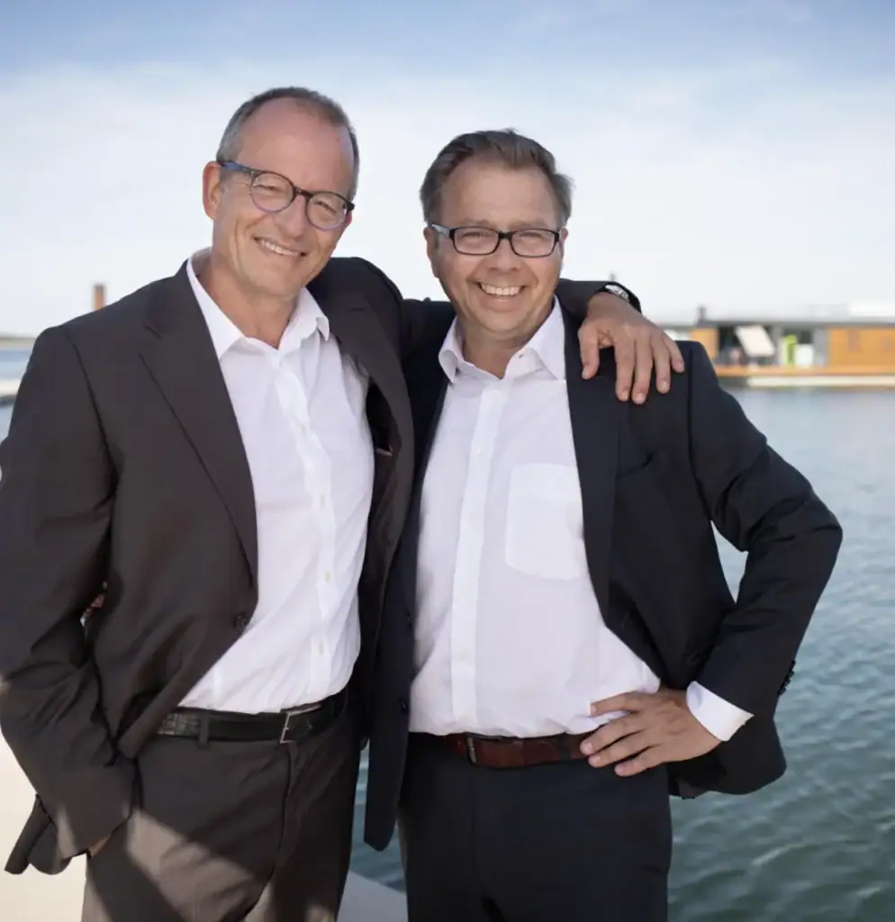 Zwei Männer in Anzügen am Wasser mit Eco Lodges im Hintergrund.